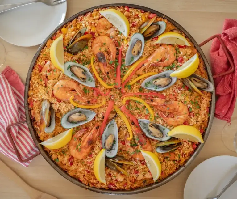 Colorful paella topped with seafood, lemon slices, and red peppers, presented on a wooden table with cutlery and a red napkin, perfect for a family meal or gathering with friends comfort food recipes